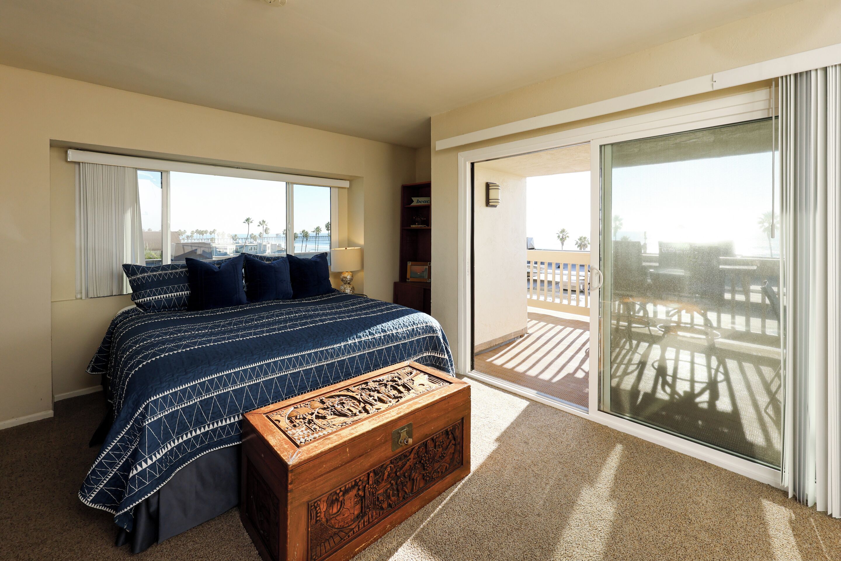 A bedroom with a bed, a wooden chest, a sliding glass door leading to a balcony, and a view of the beach.