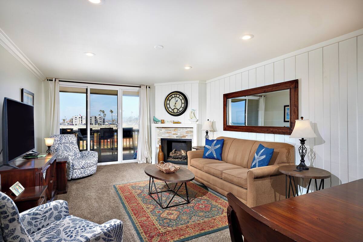 A living room with a sofa, armchairs, a coffee table, a TV, a fireplace, and a large window with a view of buildings.
