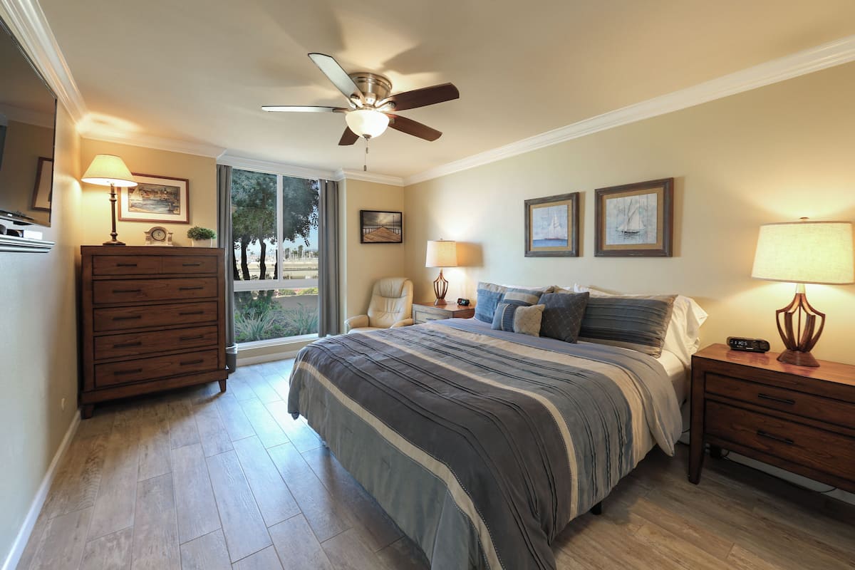 A bedroom with a bed, nightstands, a dresser, a ceiling fan, and a window with a view of trees.