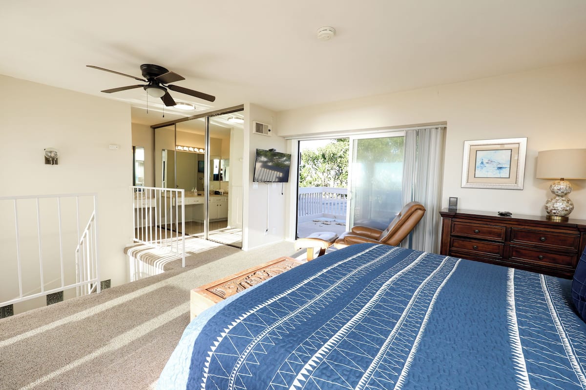 A bedroom with a large bed, a ceiling fan, a sliding glass door to a balcony, a dresser, and a TV.