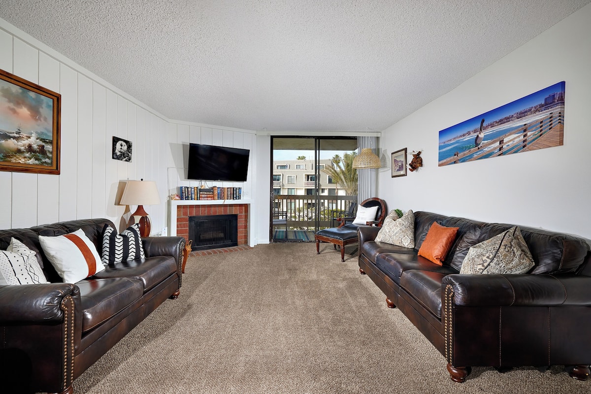A living room with two leather sofas, a flat-screen TV, a fireplace, and a large window with a view of buildings and palm trees.