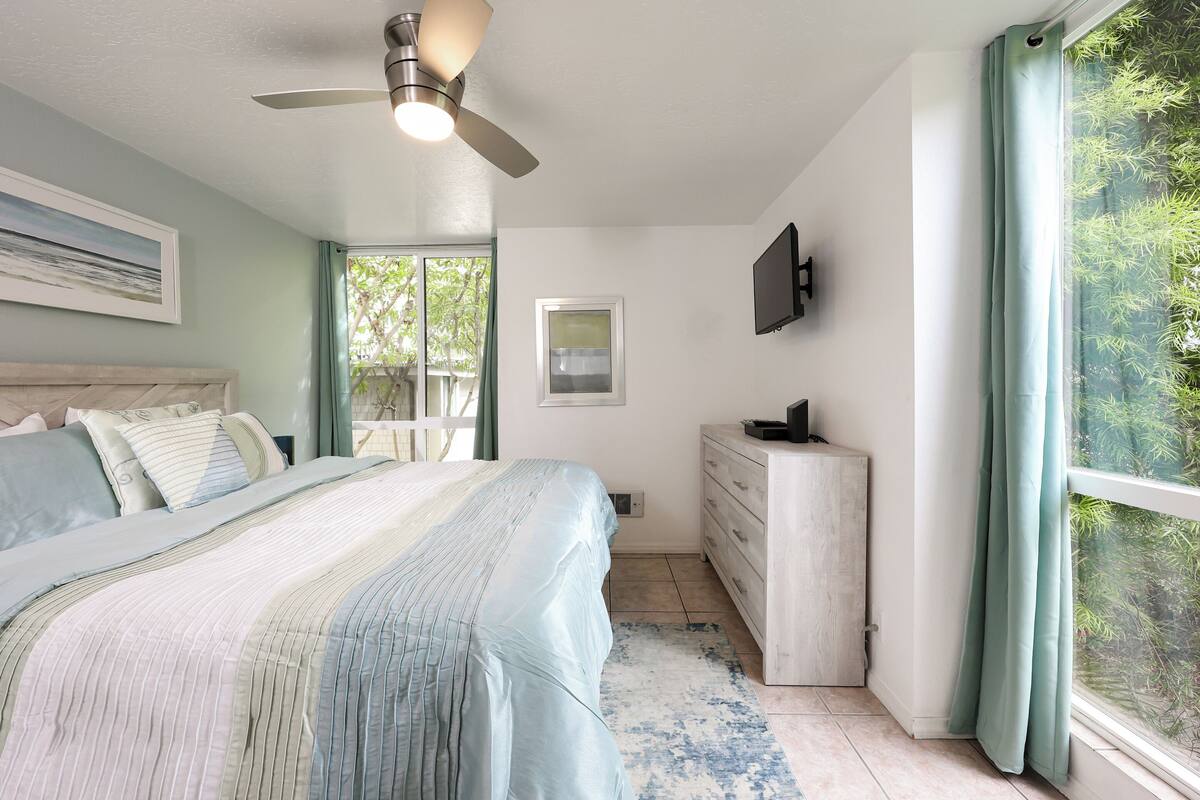 Un dormitorio con cama, cómoda, ventilador de techo, televisor y ventana con cortinas.