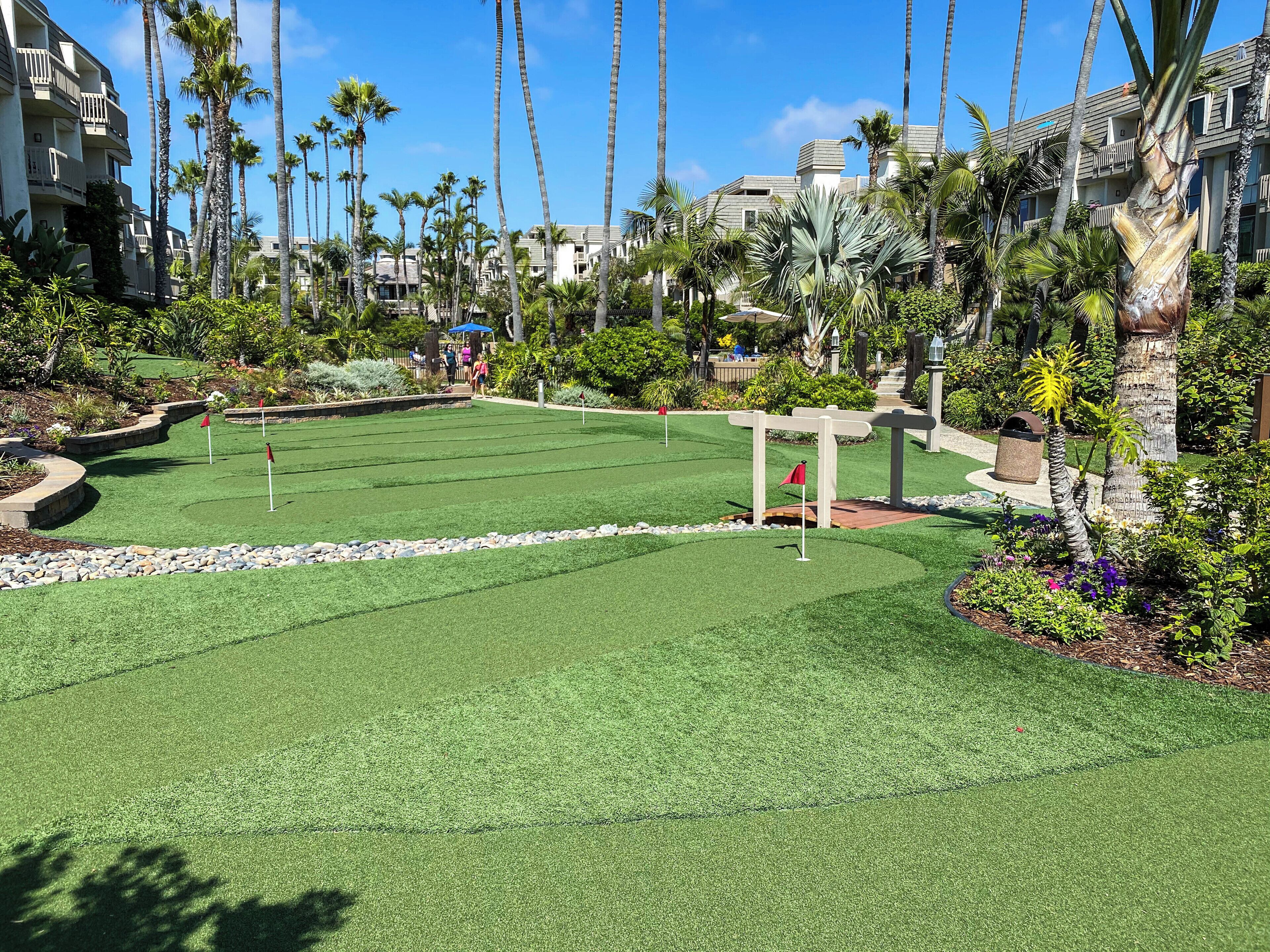 A well-maintained golf course with a clear path, surrounded by lush greenery and palm trees, with a building in the background.