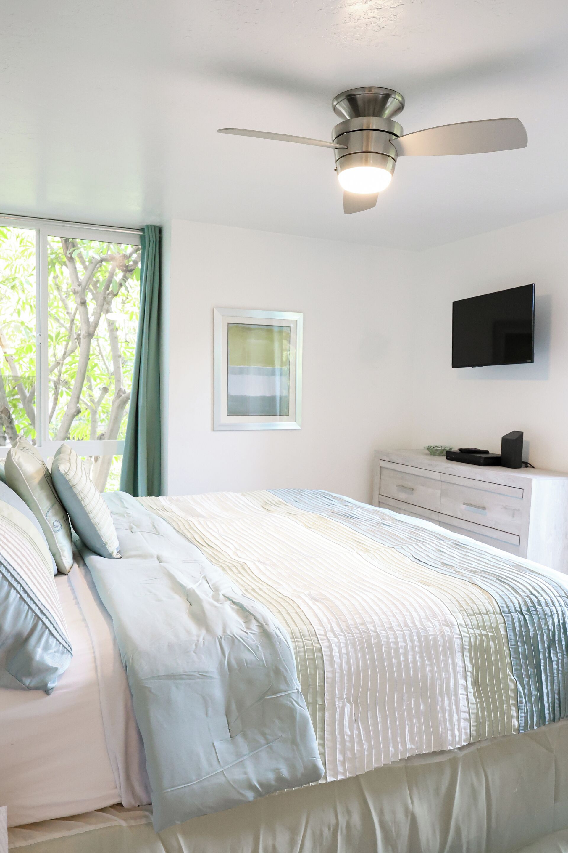 Un dormitorio con una cama, ventilador de techo, televisión y cómoda.