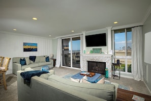 A living room with a sofa, a fireplace, a television, and a view of the beach.