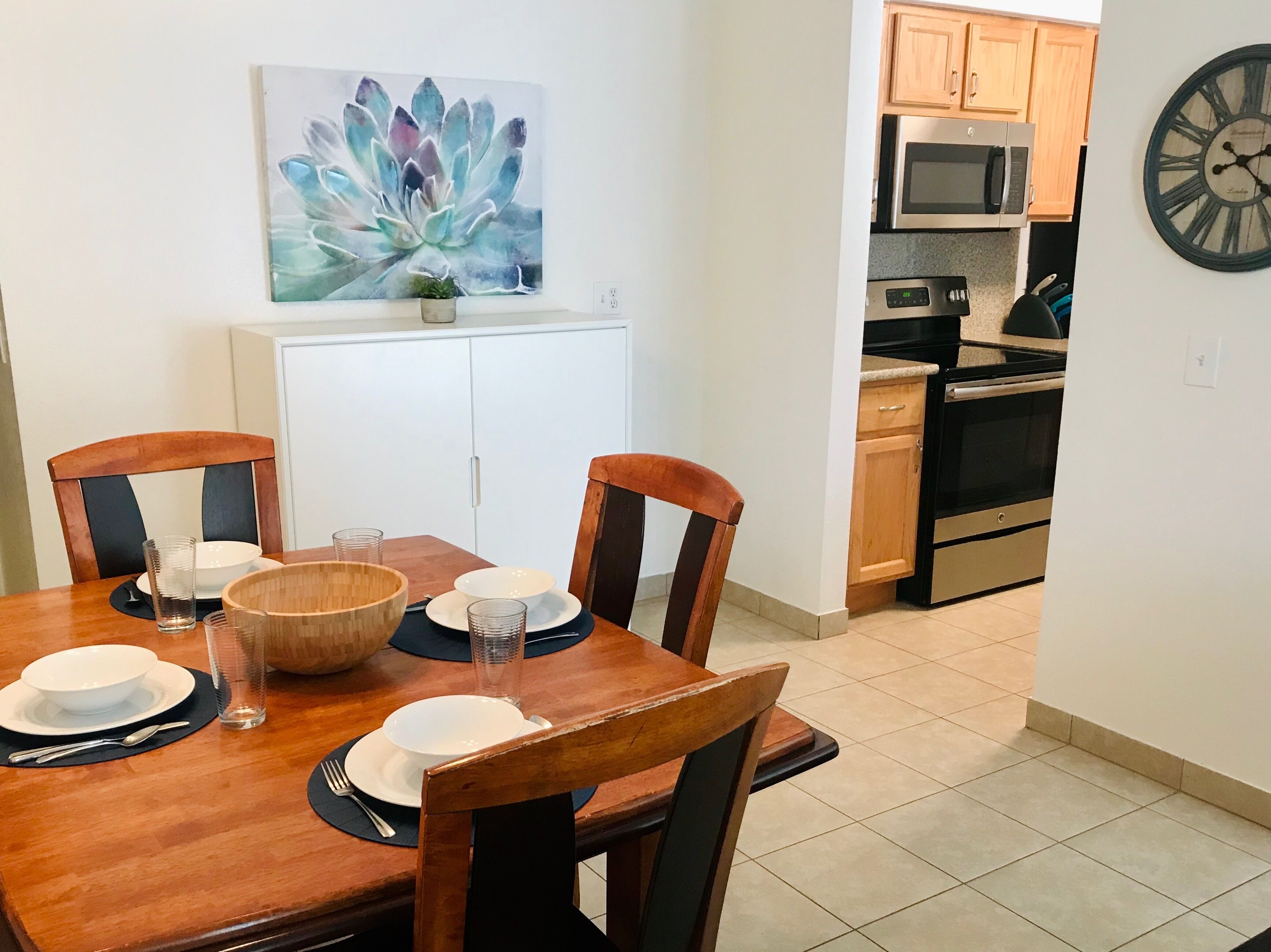 A dining area with a wooden table set for four, including plates, bowls, and glasses. A large succulent plant is on the wall above the table.