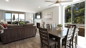 A modern living room with a sofa, dining table, and chairs. There is a TV mounted on the wall and a ceiling fan above the table. - Oceanside Beach Condos (Oceanside)