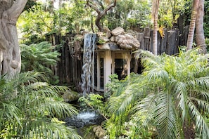Une cascade d’eau qui dévale des rochers pour se jeter dans un petit étang, le tout entouré d’une végétation luxuriante et d’une clôture en bois.