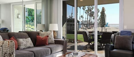 A modern living room with a grey sofa, brown ottoman, and a black chair. A glass door leads to a balcony with outdoor furniture and palm trees.
