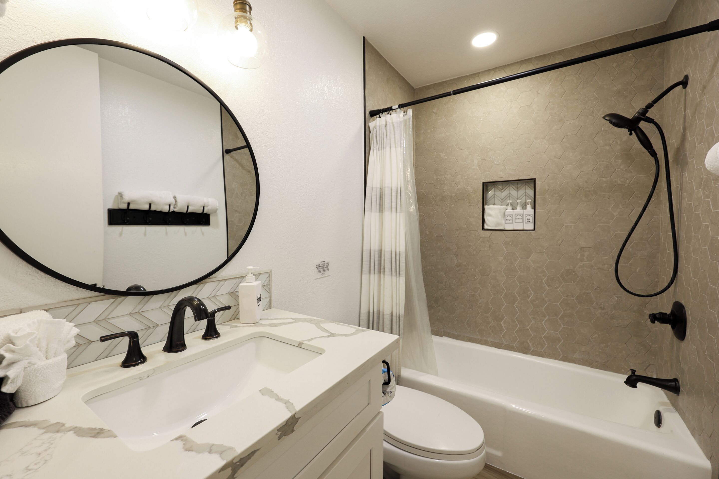 A bathroom with a round mirror, a sink with a marble countertop, a toilet, and a bathtub with a showerhead.