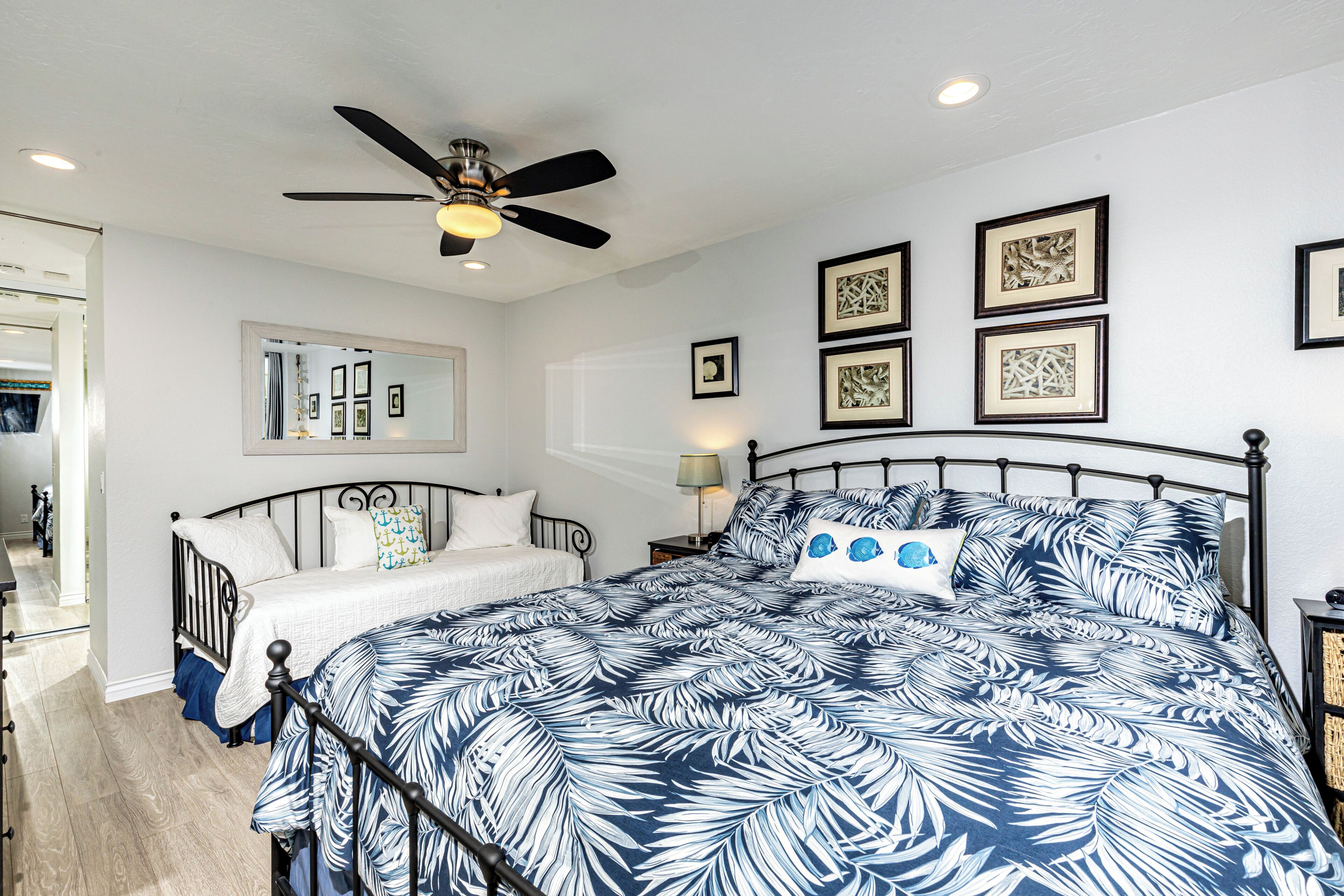 A bedroom with a bed, a ceiling fan, a mirror, and framed artwork on the wall.