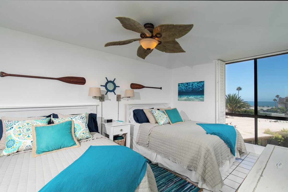 A hotel room with two beds, a ceiling fan, a view of the beach, and a starfish artwork on the wall.
