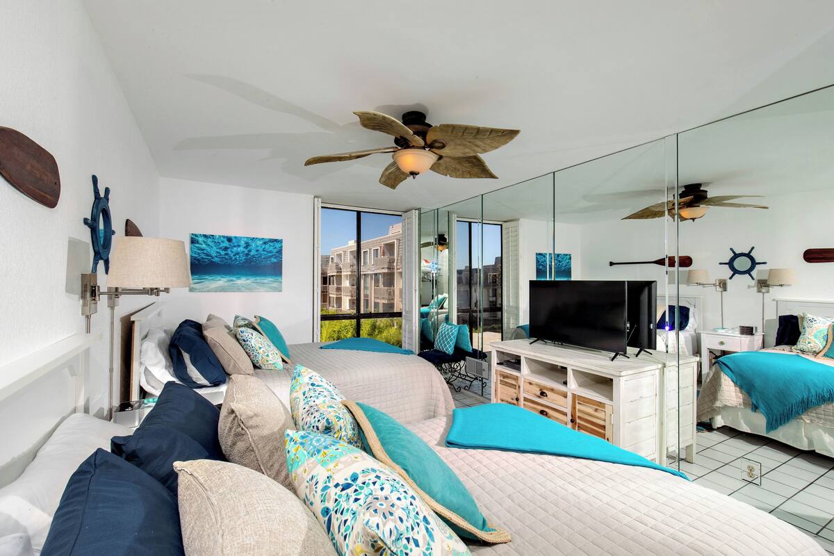 A modern bedroom with a large bed, a TV, a desk, and a balcony with a view of buildings.