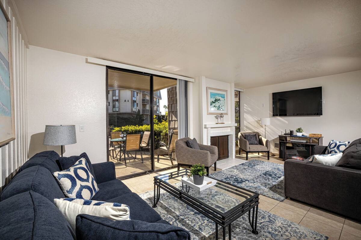 A modern living room with a sofa, armchairs, a coffee table, and a TV. There is a sliding glass door leading to an outdoor area with a table and chairs.
