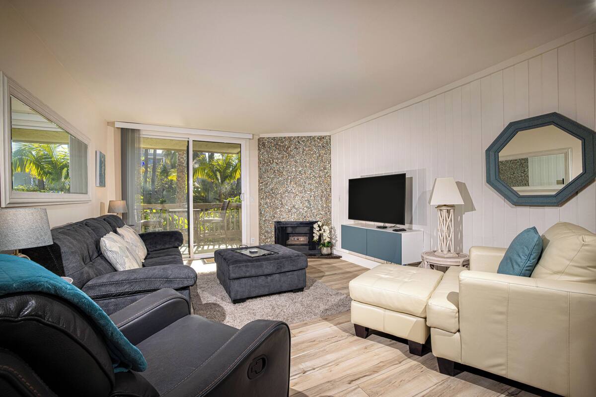 A modern living room with a fireplace, a flat-screen TV, and a balcony with a view of palm trees.