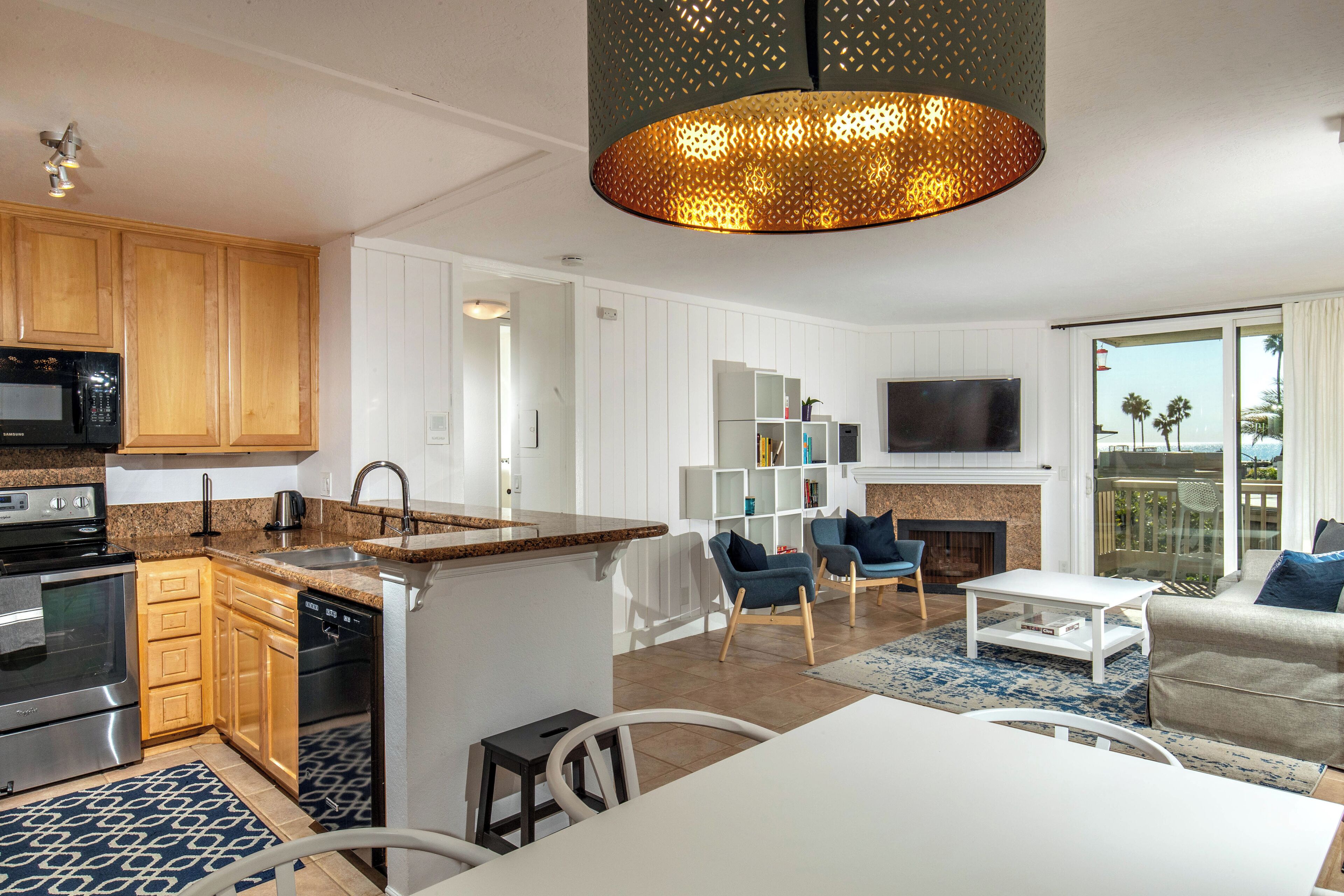 Une cuisine moderne avec des meubles en bois, un four à micro-ondes et un lave-vaisselle. On y trouve un coin repas avec une table et des chaises blanches, ainsi qu’un salon avec un canapé et un téléviseur.