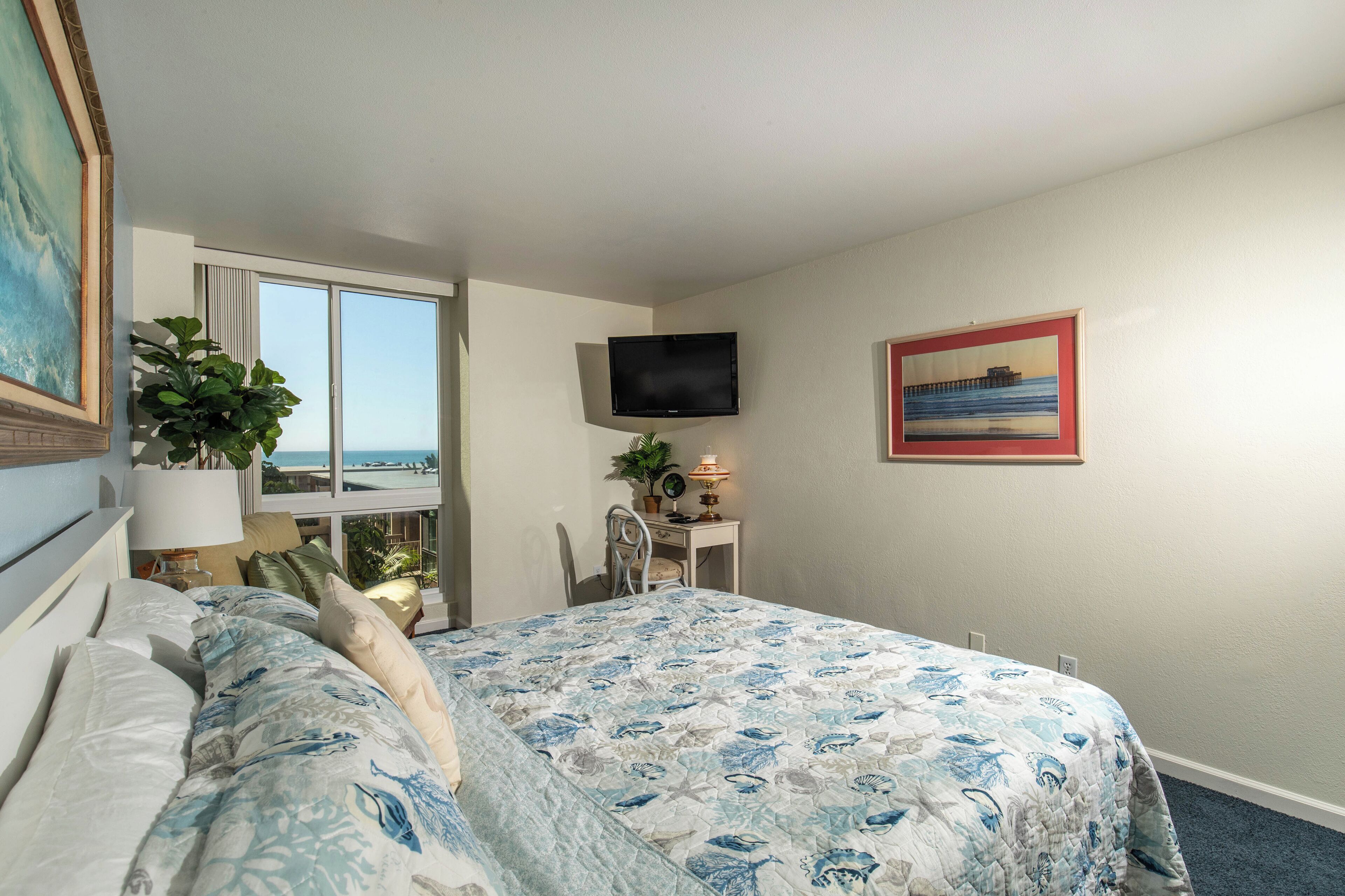 A bedroom with a bed, a desk, a chair, a TV, a window, and a plant.
