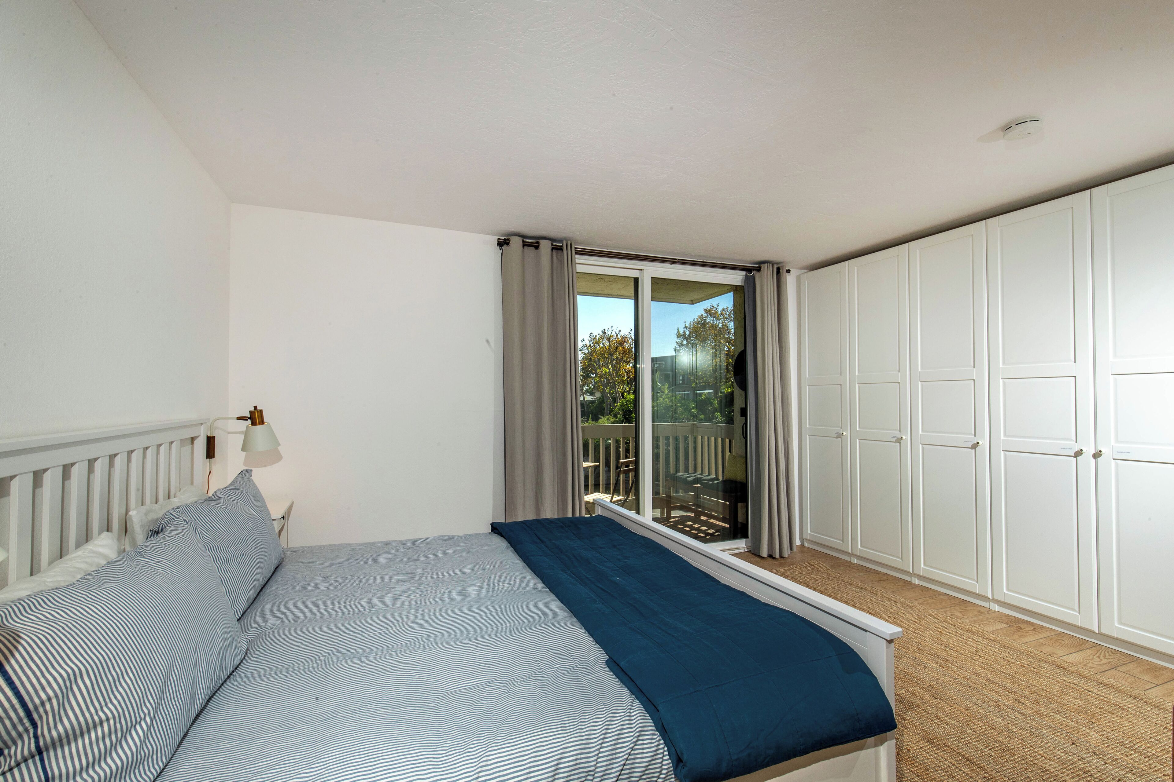 Ein Schlafzimmer mit Bett, Nachttisch, Lampe, einem Fenster mit Vorhängen und einem Kleiderschrank.