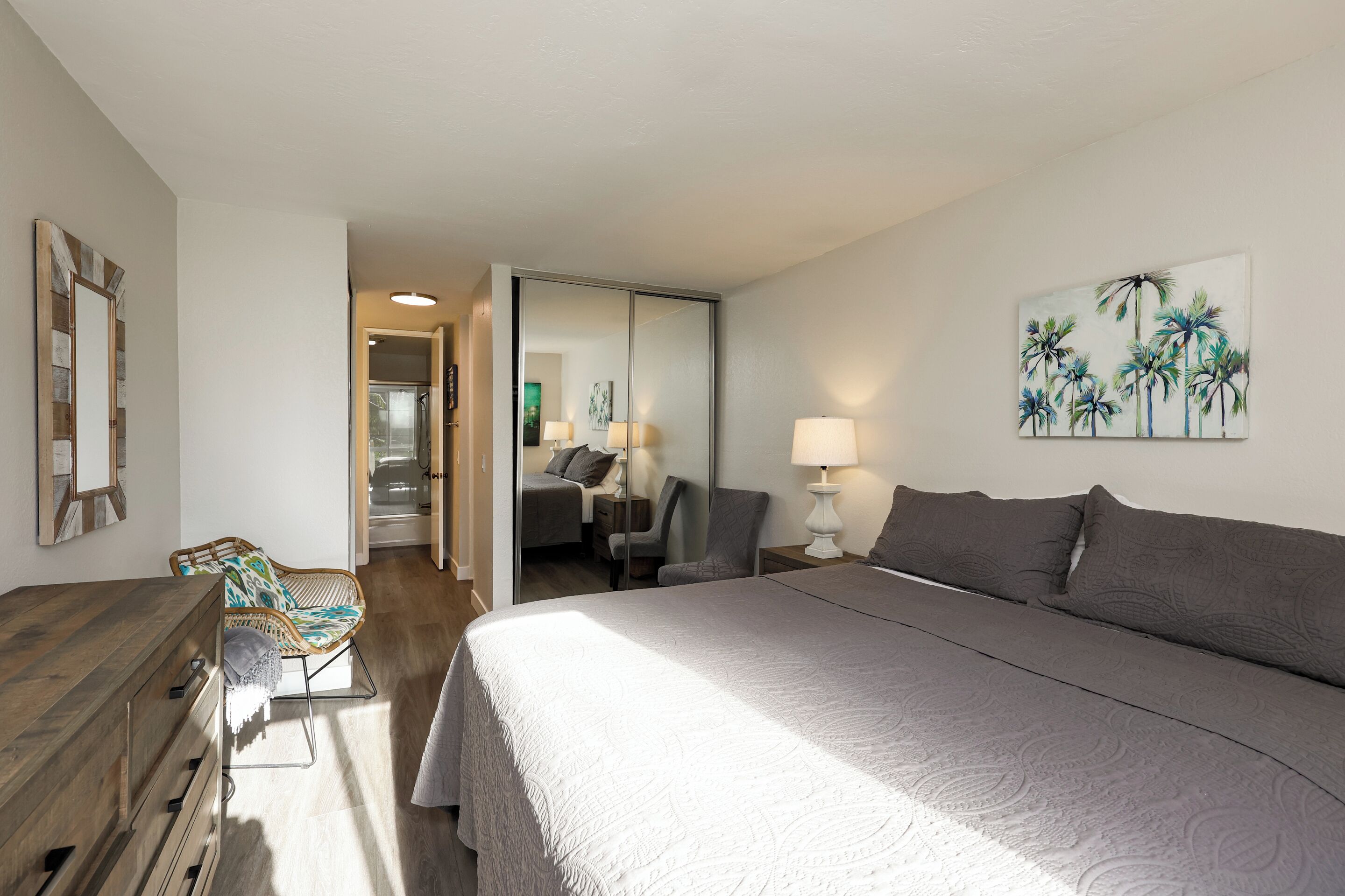 Quarto com uma cama grande, penteadeira de madeira, cadeira de vime e um quadro com palmeiras.