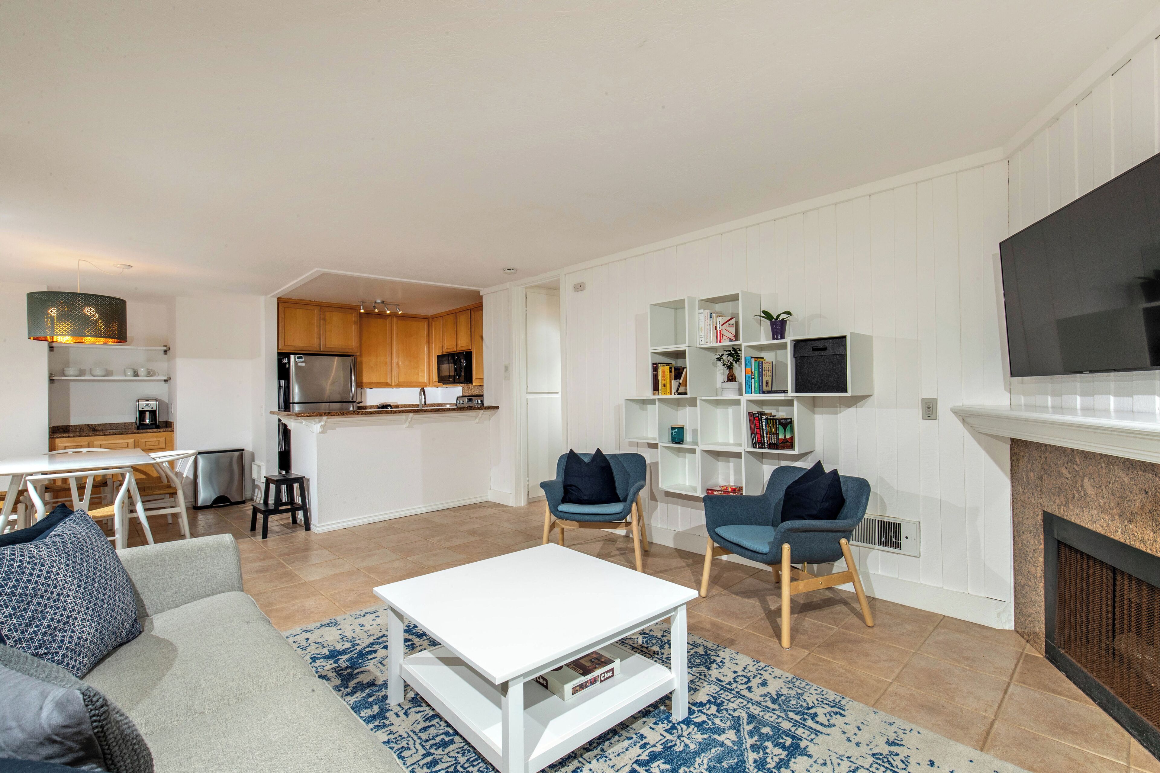 Un salon moderne avec un canapé blanc, une table basse et deux fauteuils bleus. On y trouve également un coin cuisine avec des meubles en bois et un micro-ondes encastré. Un téléviseur à écran plat est fixé au mur, au-dessus d'une cheminée.