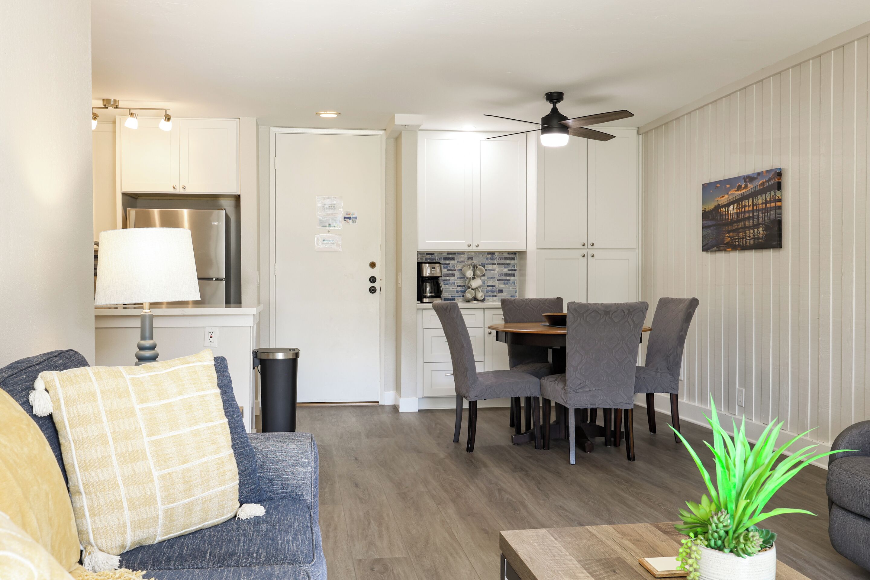 A modern living room with a sofa, a dining table with chairs, a ceiling fan, and a kitchen area with cabinets and a refrigerator.