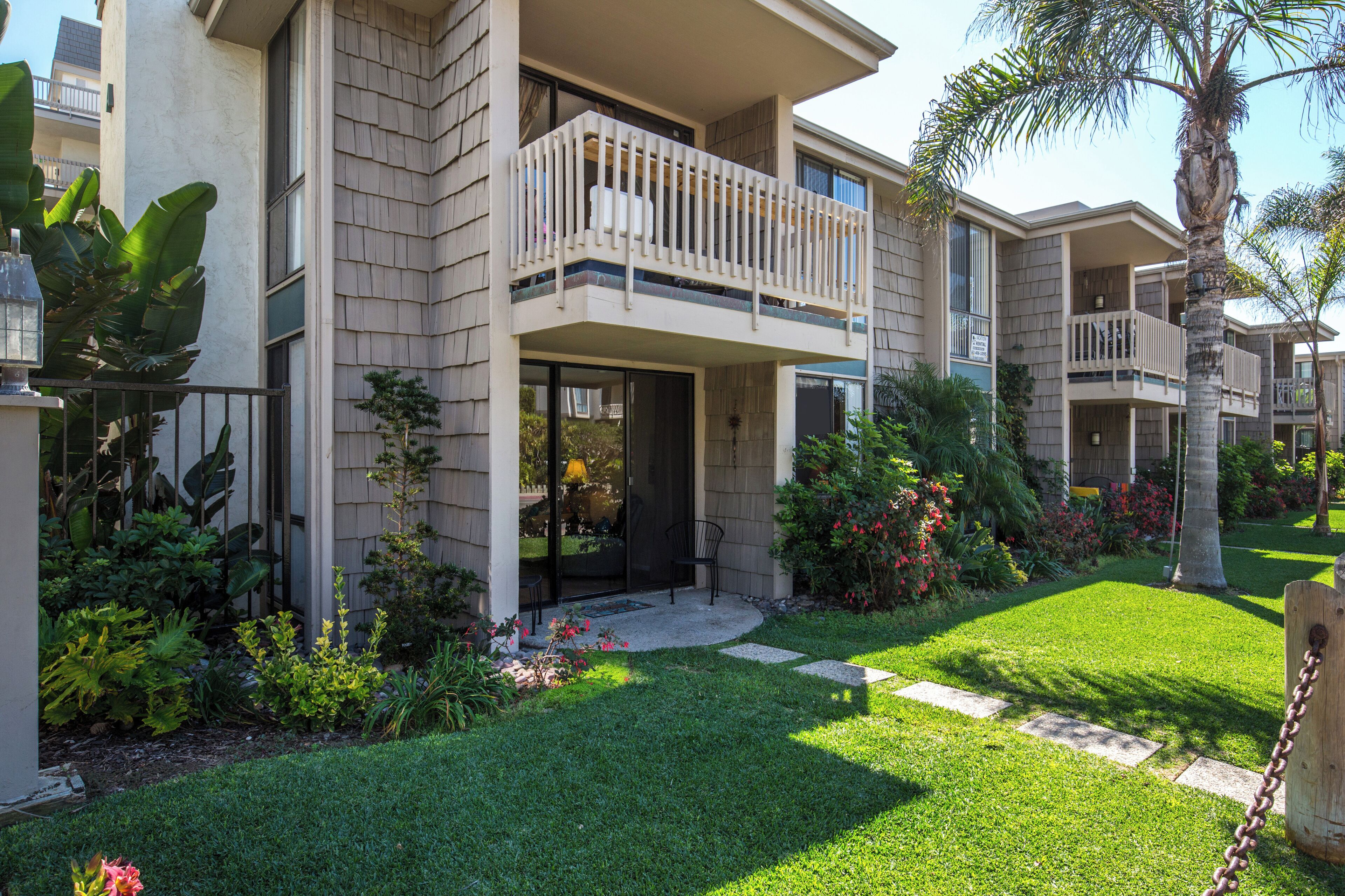 A modern multi-story residential building with a balcony, a well-maintained lawn, and various plants.