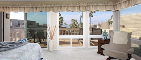 A bedroom with a bed, a rocking chair, a window with a view of palm trees and a balcony, and a large window with a view of the cityscape.