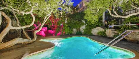 A swimming pool with a wooden deck, surrounded by lush greenery and rocks.