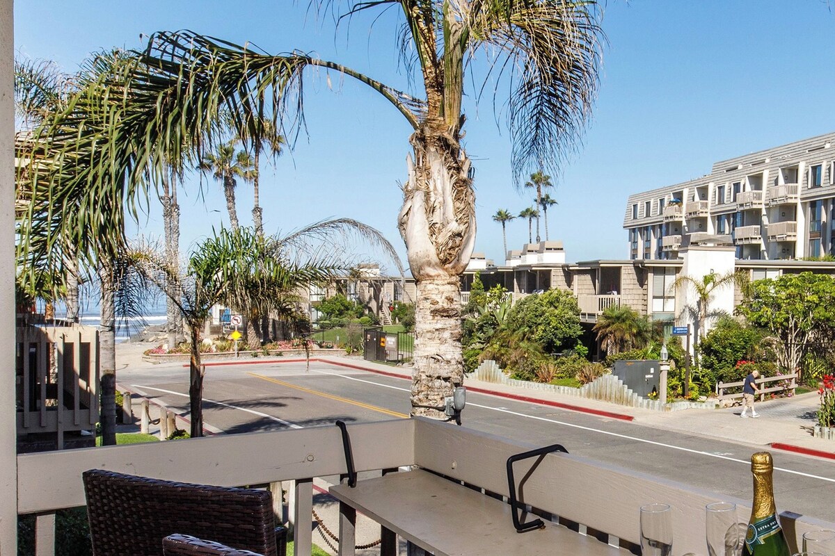 A balcony with wicker chairs and a table, overlooking a street with palm trees and a building with balconies.