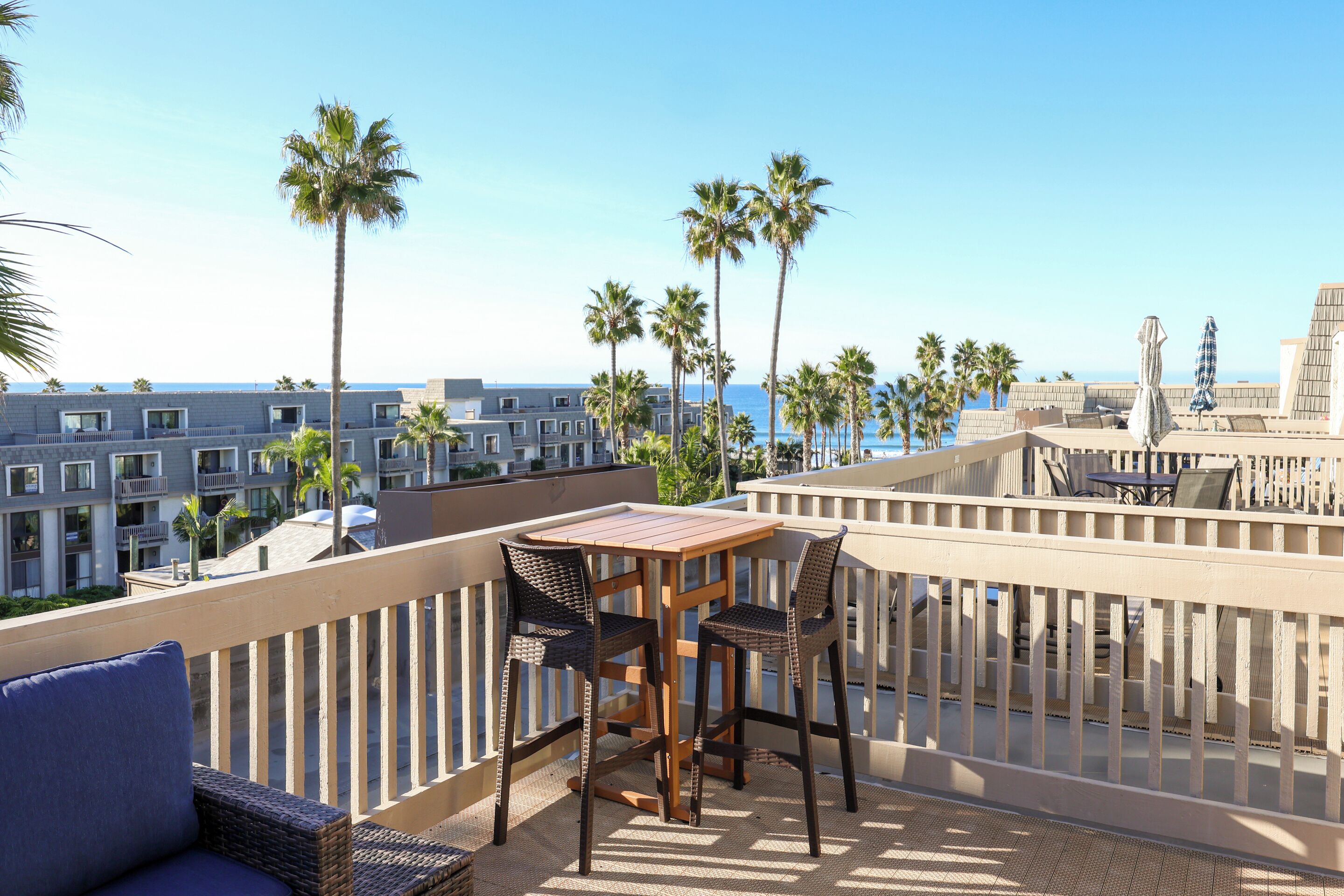 Un balcon avec une table et des chaises en bois, donnant sur une plage et sur les bâtiments d’un hôtel.