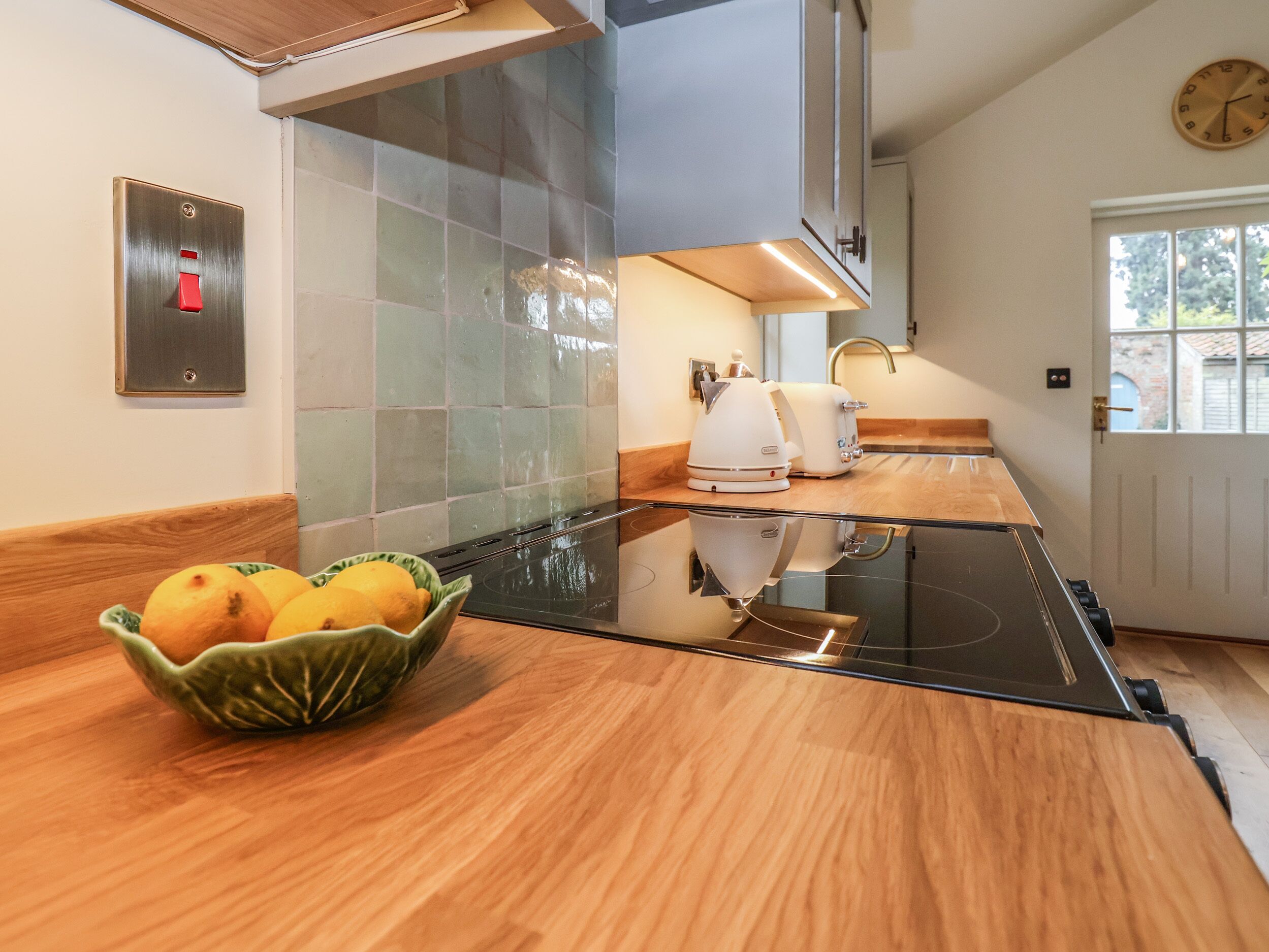 A modern kitchen with a glass cooktop, a bowl of fruit, a toaster, and a clock on the wall.