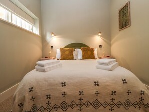 A neatly made bed with white linens and a patterned blanket, two bedside tables with lamps, and a window with blinds. - Rookery Cottage (Saxmundham)
