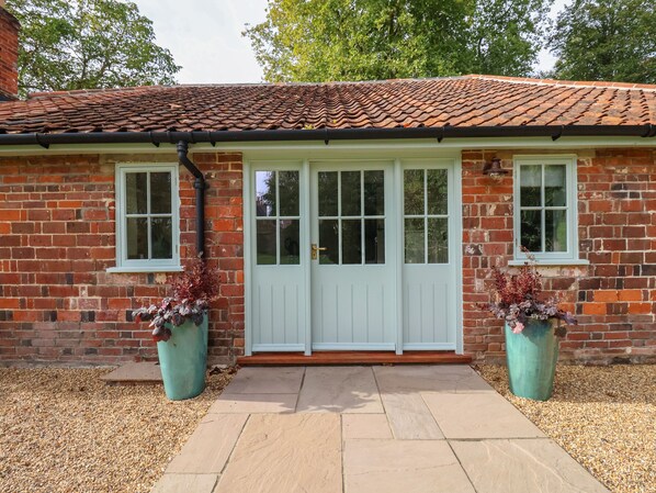 A brick building with a tiled roof, a white door with glass panels, and two windows. There are two large planters with dried plants in front of the door. - Rookery Cottage (Saxmundham)