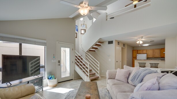 A spacious living room with a large TV, a sofa, a coffee table, and a staircase leading to a kitchen area. - Tranquil & bright loft-style townhouse near Sabino Canyon!   (Tucson)