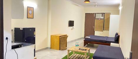 A compact hotel room with a kitchenette, two single beds, a small wooden table with water bottles, a TV, and a ceiling fan.