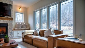 A cozy living room with a stone fireplace, leather sofa, and large windows offering a view of a wooded area. - New Farmhouse-Hot Tub, King Beds, Game Room, Pro Golf Sim & Amazing Valley View! (Montgomery)