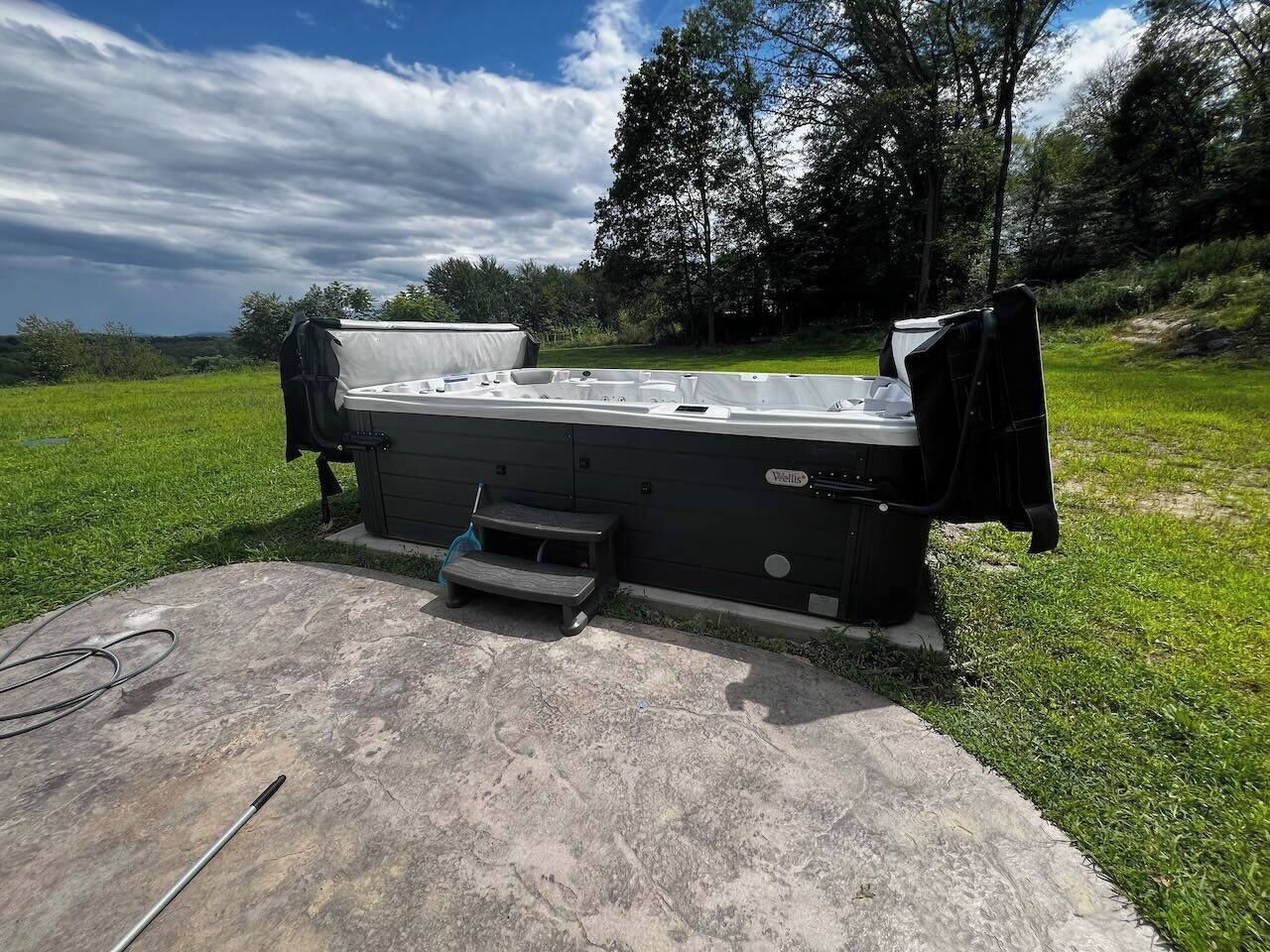 A hot tub with a cover, a small step stool, and a hose on a concrete surface in a grassy area.