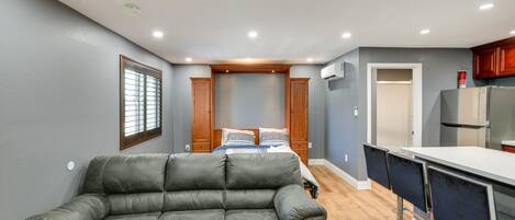 A compact living space with a sofa, a bed, a kitchenette, and a window with blinds.