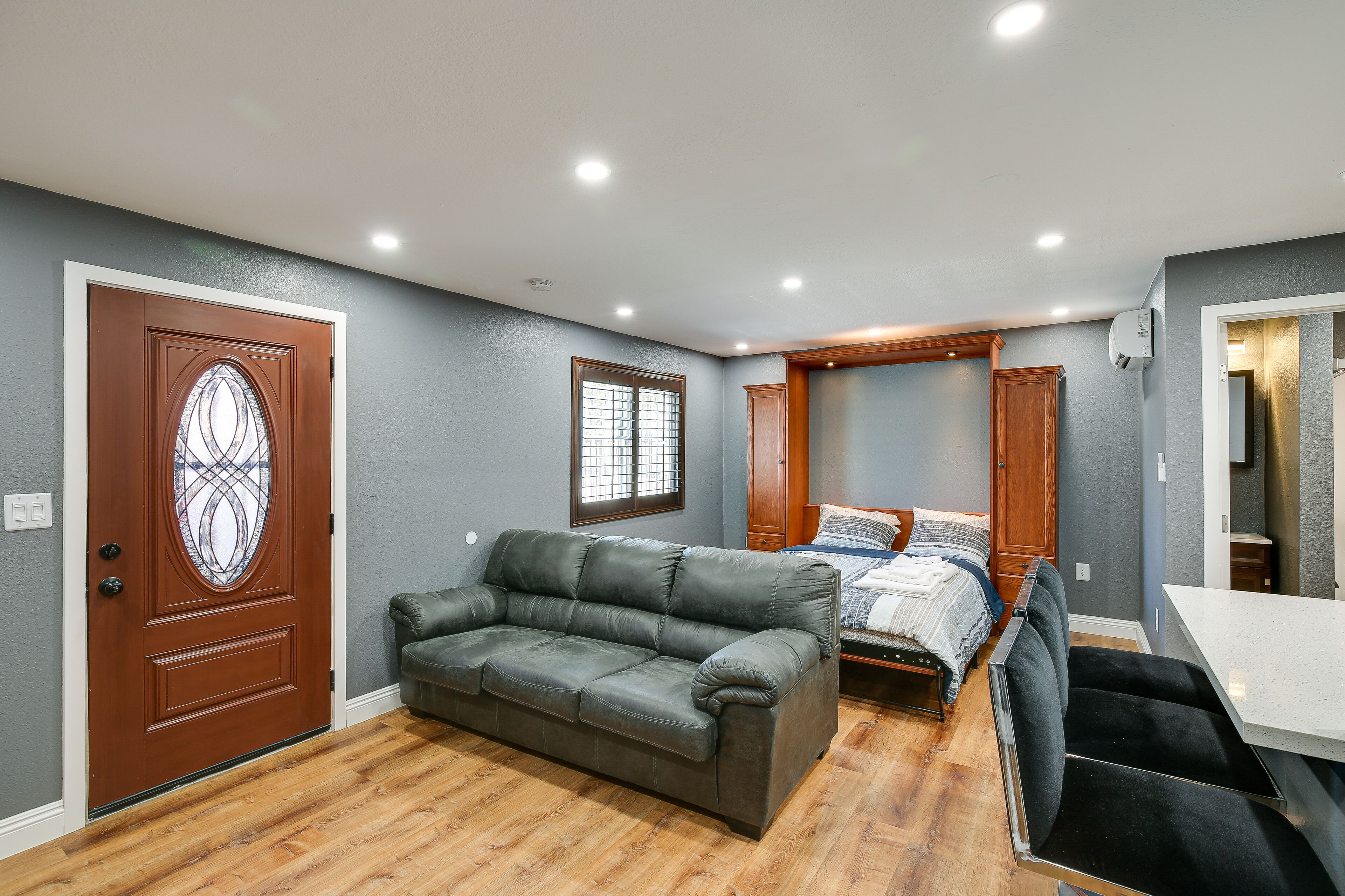 A compact bedroom with a wooden door, a grey leather sofa, a bed with a wooden headboard, and a small table.