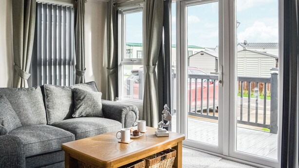 A living room with a grey sofa, a wooden coffee table with a mug and a small sculpture, and large sliding glass doors leading to a balcony. - Patrington Haven is a pristine lodge park with an array of facilities. (Patrington, near Hull)
