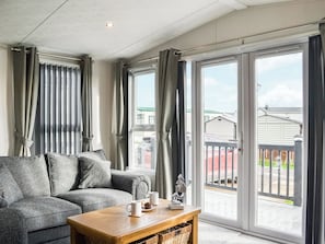 A living room with a grey sofa, a wooden coffee table with a mug and a small sculpture, and large sliding glass doors leading to a balcony.