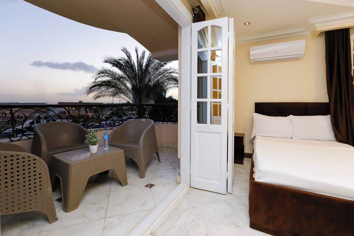A balcony with wicker furniture, a bed with white linens, and an air conditioning unit.