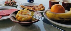 Une table est dressée avec des assiettes remplies de nourriture, notamment un bol de poulet frit, une banane et une part de pizza. On y trouve également des tasses à café et une assiette avec une salade.