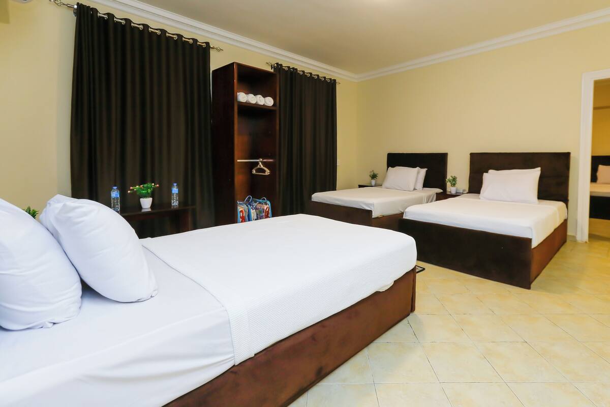 A hotel room with two single beds, a large bed, a wardrobe, a shelf with toiletries, and a potted plant.