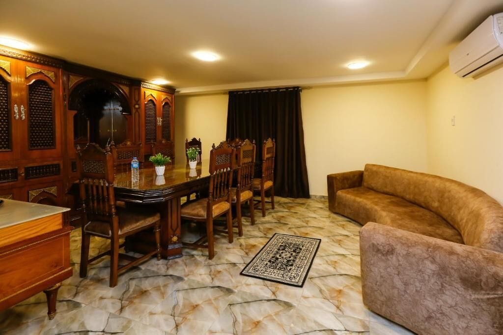 A dining area with a wooden table and chairs, a sofa, and a patterned rug on a marble floor.