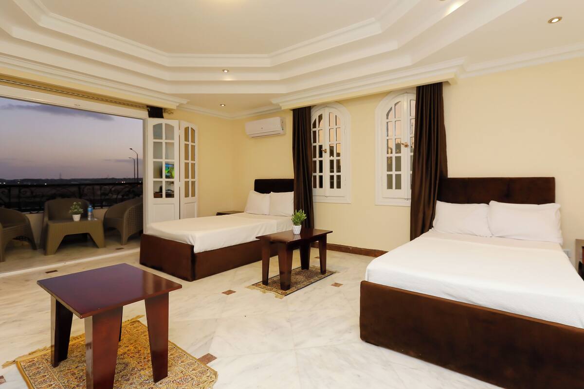A hotel room with a large bed, two smaller beds, a balcony with chairs, a small table, and a view of the cityscape.