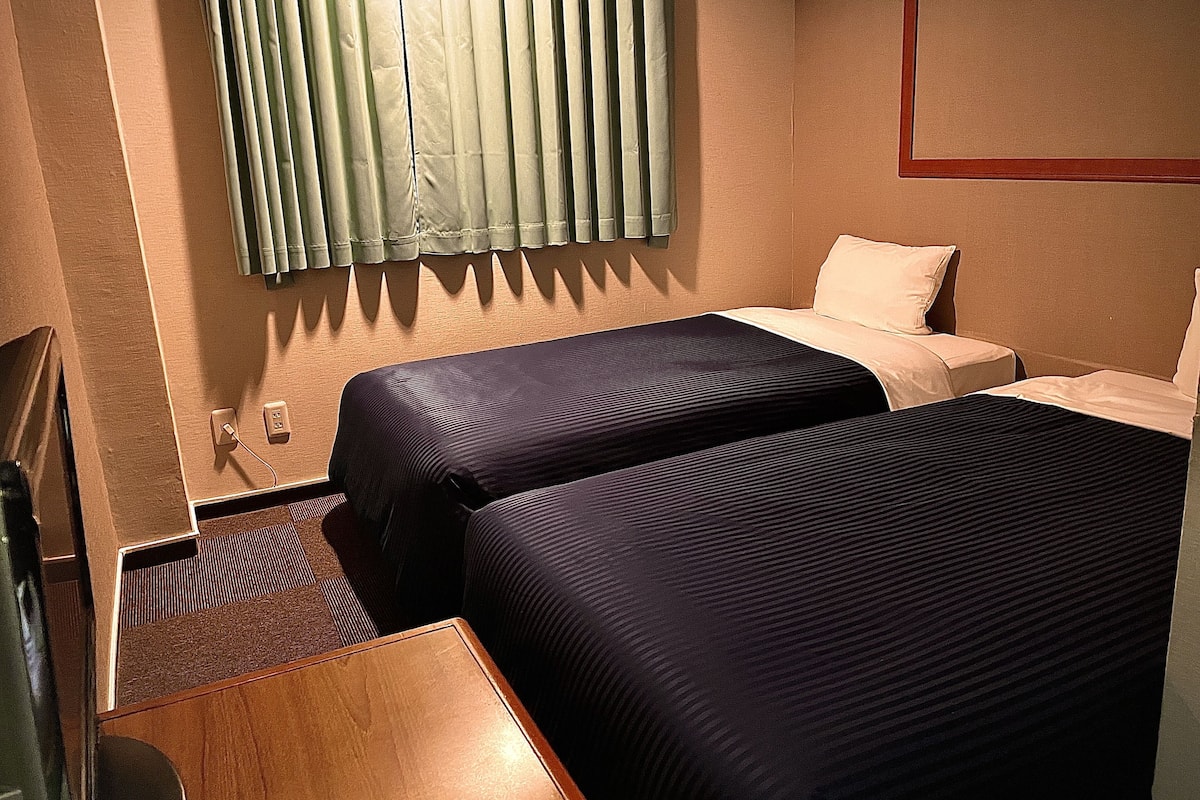 Une chambre d’hôtel avec deux lits, un bureau en bois, une fenêtre avec des rideaux et un téléviseur fixé au mur.
