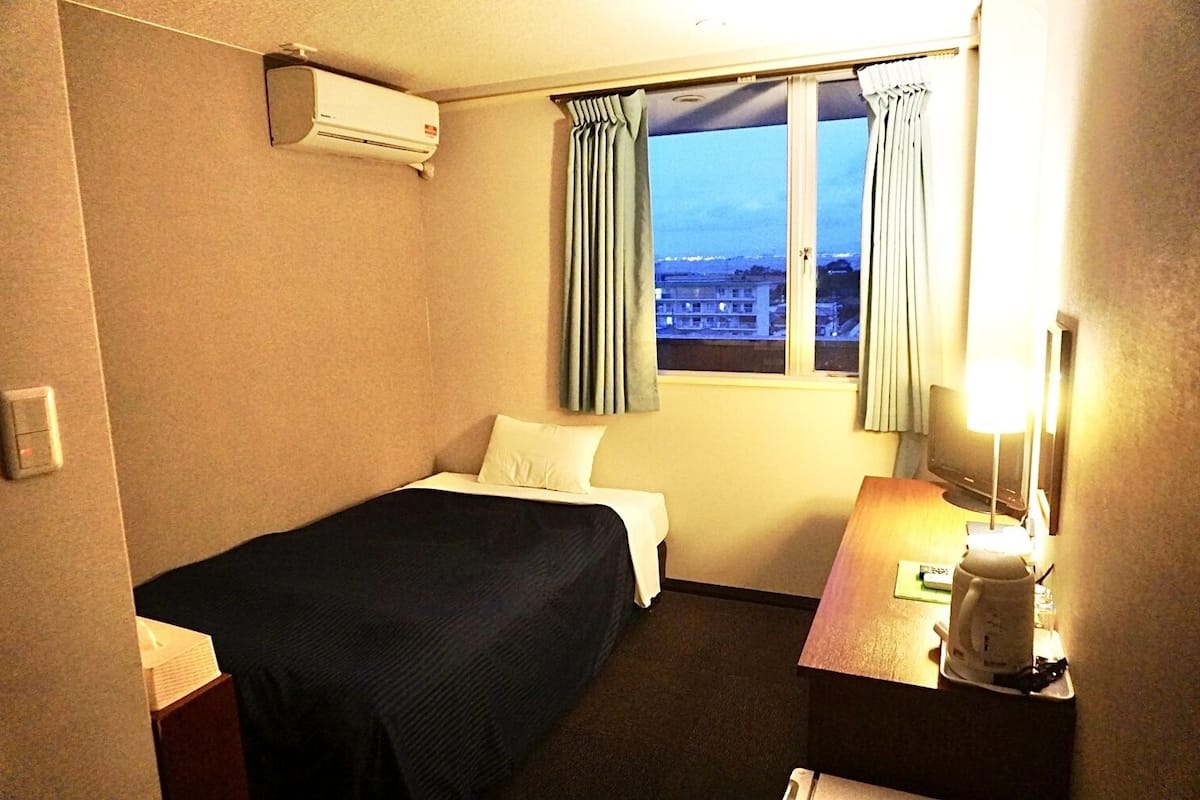 Une chambre d’hôtel avec un lit, un bureau avec une lampe, un climatiseur et une fenêtre avec des rideaux.