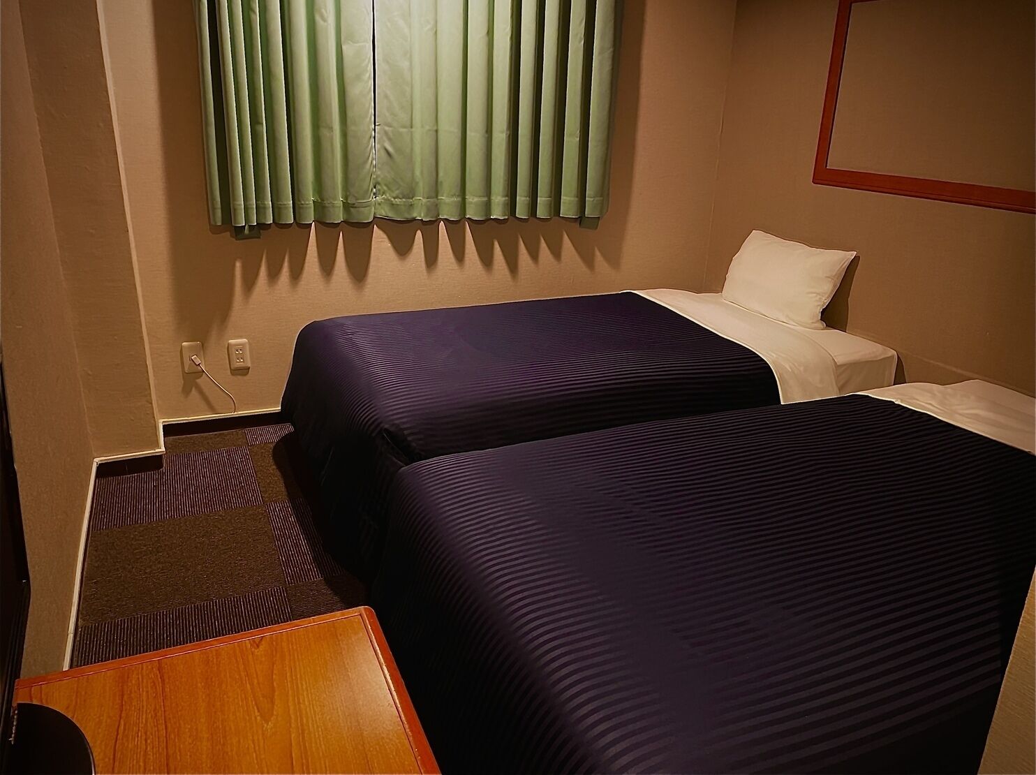Quarto de hotel com duas camas, uma mesinha, uma cadeira e uma janela com cortinas.