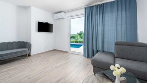 Ein modernes Wohnzimmer mit einem grauen Sofa, einem wandmontierten Flachbildfernseher und Blick auf einen Swimmingpool durch eine Glastür.